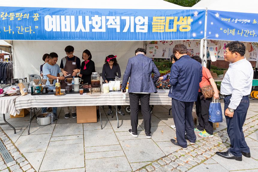 들다방 야외 천막 부스 풍경. "노란들판의 꿈, 나, 간다"라는 슬로건과 함께 '예비사회적기업 들다방’이라 쓰인 현수막이 걸려 있다. 그 아래 기다란 테이블이 펼쳐져 있고, 일꾼들로 보이는 네다섯 명의 사람들이 분주하다. 부스 바깥으로 기다리는 이들이 긴 줄이 늘어서 있다.