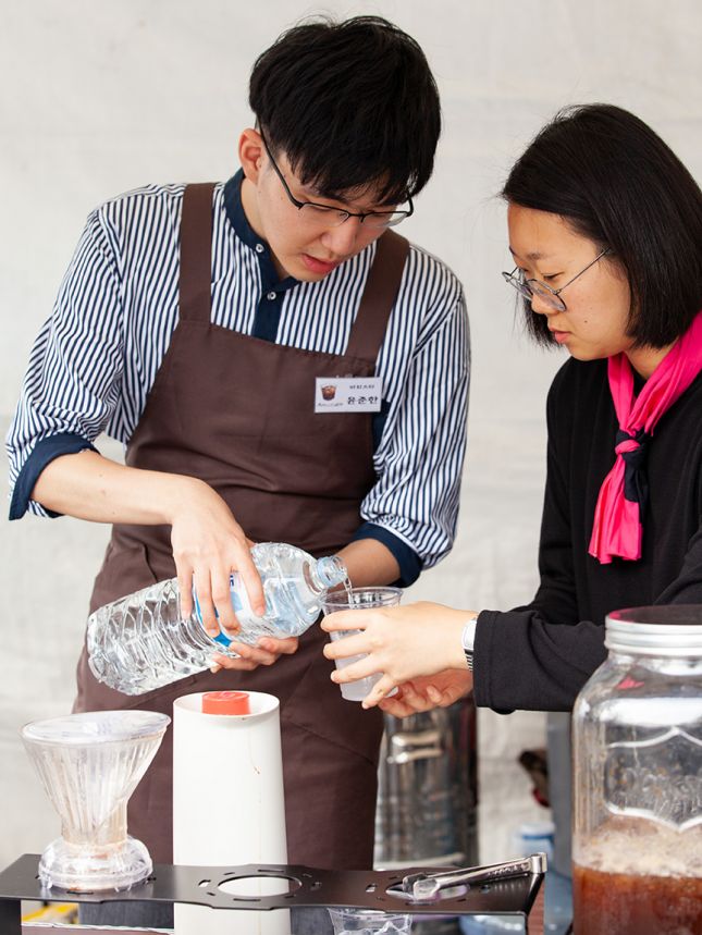 야외 천막 부스 안에서 앞치마에 바리스타 명찰을 단 남성이 양 손으로 생수통을 들고 물을 따르는 모습. 옆에 한 여성이 얼음이 담긴 테이크아웃컵을 받치며 조력하고 있다. 이들 앞으로 아이스 아메리카노가 담긴 커다란 유리 디스펜서와 핸드드립 전용 받침대와 드리퍼 등이 보인다. 