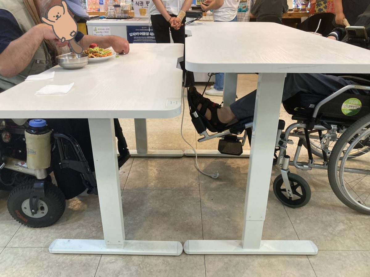 들다방 카페 홀에서 두 사람이 테이블을 가운데 두고 앉아 있는 모습 측면 사진. 마주 놓인 두 테이블 높이가 15센티미터 정도 차이가 난다. 왼쪽 테이블에는 전동휠체어 사용자, 오른쪽 테이블에는 수동 휠체어 사용자가 저마다 높이를 다르게 조정해 이용 중이다. 그들 뒤로 배식을 위해 사람들이 줄을 서거나, 카페를 이용 중이다.