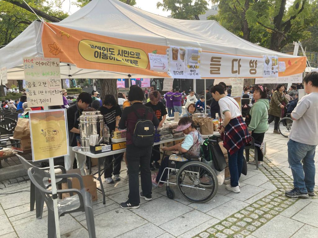 마로니에공원에 배치된 들다방 부스 측면 사진. 테이블을 사이에 두고 손님, 스태프로 부스 안이 북적인다. 부스 상단에는 들다방이라 크게 인쇄된 천막이 가로로 걸려 있다.