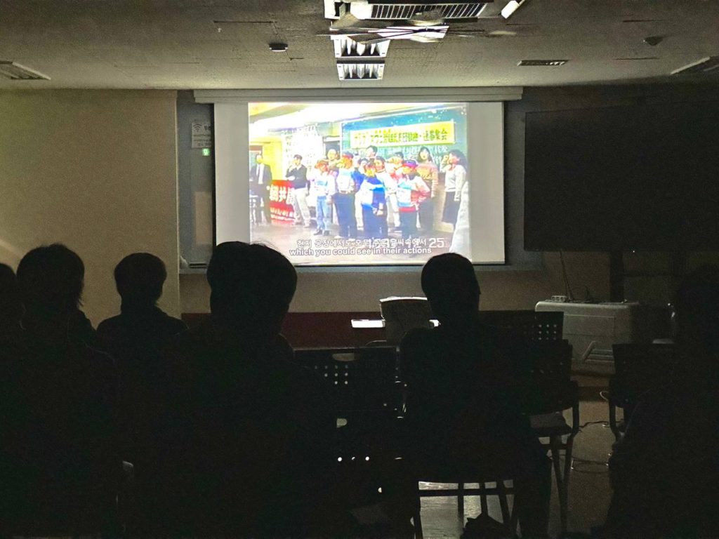 어두운 공간에 은막만이 환하다. 스크린에 띄워진 영상 속에 열 명 가까이 빨간 머리띠를 두른 이들이 구호가 적힌 현수막 앞에서 열을 갖춰 주먹을 치켜 들거나 서서 구호를 외치는 모습. 그 모습을 지켜보는 관객의 뒷모습이 뒤통수만 어두운 그림자로 보인다.