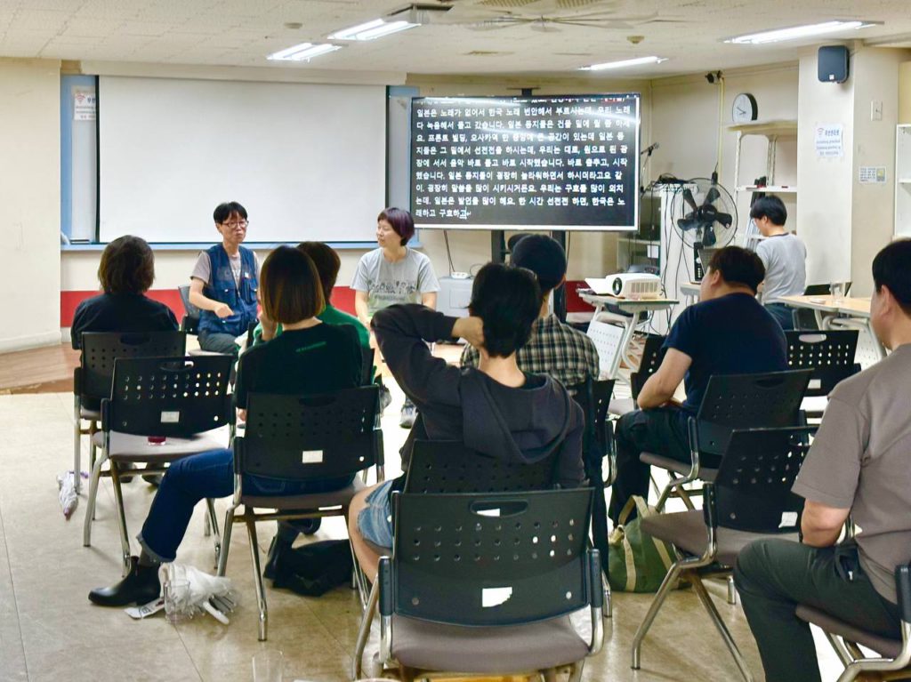 스크린 앞 무대에 초대손님과 진행자가 앉아 있고, 그 뒤로 관람객 객석에 사람들이 저마다 앉아 무대를 바라보고 있다. 무대 앞 오른편에 큰 스크린으로 문자통역 화면이 제공되고, 문자 통역 속기하는 사람 뒷모습도 보인다.
