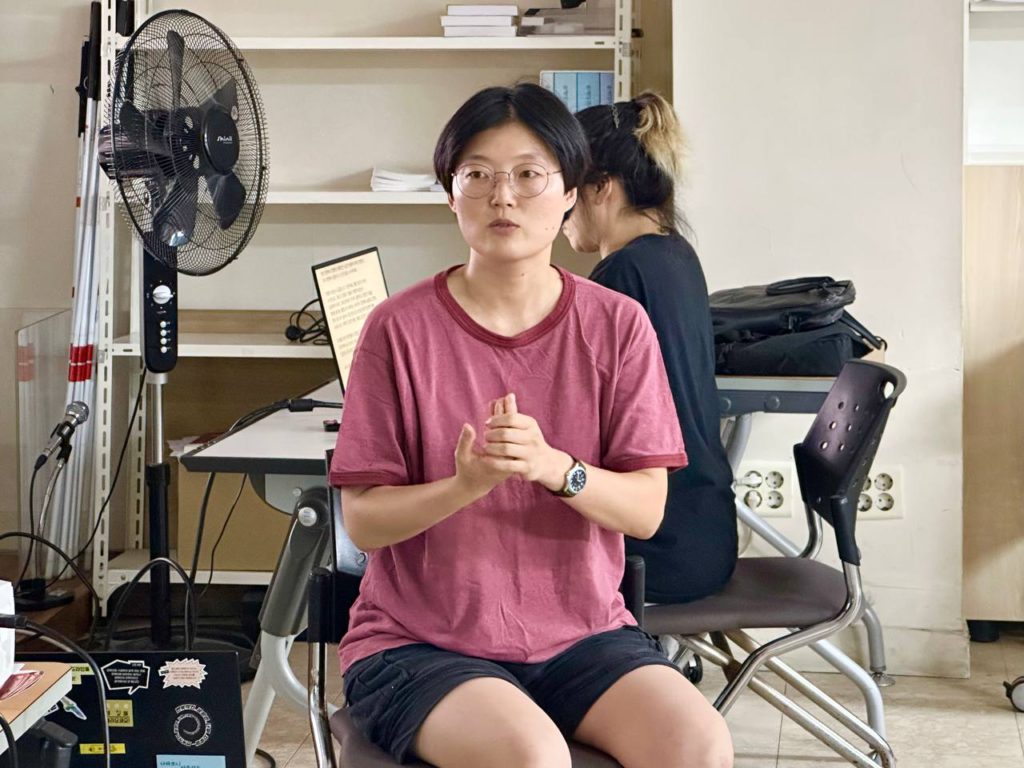 의자에 앉아 무언가 말하고 있는 진행자 사진. 뒤쪽에는 문자통역을 위해 속기중인 사람과 노트북이 보인다.