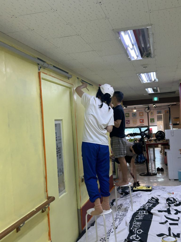 들다방 복도 페인트칠 사진. 바닥에 버려진 천막을 한껏 펼쳐져 있고, 비닐도 깔려 있다. 저마다 페인트 롤러나 붓을 든 사람들 셋이 간이 의자 위에 올라 서서 벽면 구석구석 붓칠을 하고 있다. 