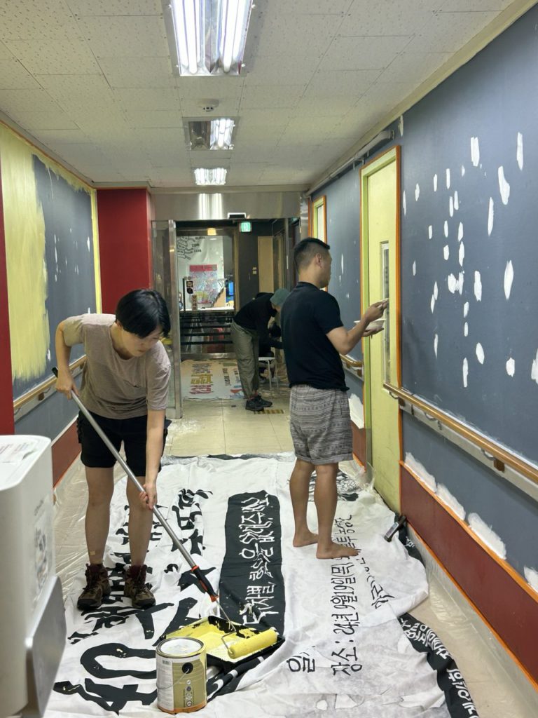 들다방 내부에서 출구 쪽을 바라보며 찍은 복도 사진. 왼쪽 벽면에 레몬색 페인트를 칠하려 롤러에 물감을 적시려 허리를 구부린 사람. 오른쪽 벽면은 아직 본격적인 페인트칠 시작을 하지 않아 남색 벽지와 하얀 벽 점토 매운 자국이 그대로다. 오른편에는 한 사람이 문을 레몬색으로 칠하고 있다. 바닥에는 폐현수막이 펼쳐져 있다. 