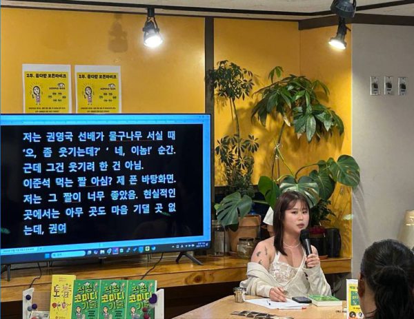 무대 앞에 책 적정 코미디 기술이 나열된 가운데 북토크 중 마이크를 들고 얘기하는 금개 작가님 사진. 옆의 모니터에는 속기 문자가 띄워져 있는데, 권영국 선배 물구나무 설 때 좀 웃기는데? 했다는 말이 띄워져 있다.