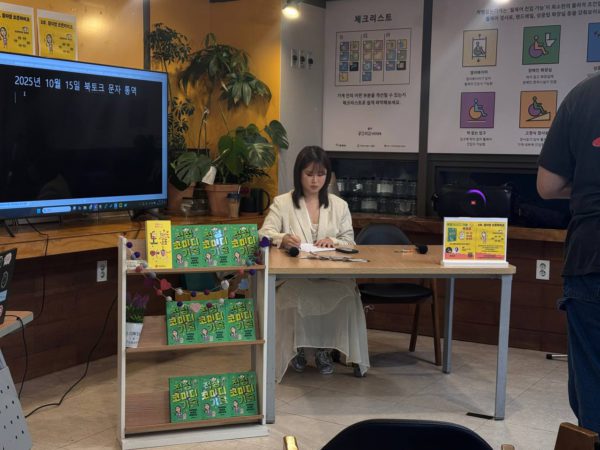 들다방을 무대로 금개님이 테이블에 앉아 싸인하는 모습, 테이블 앞 책선반에 적정코미디기술 책이 가지런히 전시돼 있다.