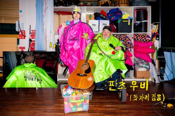 교실 한구석 다용도 수납장 앞 핑크와 초록 판초 우비를 입은 두 사람. 왼편 사람은 전면에 큰 글씨로 장애 해방과 그림이 인쇄된 우비를 입었고, 오른편 사람은 초록 우비를 입었다.