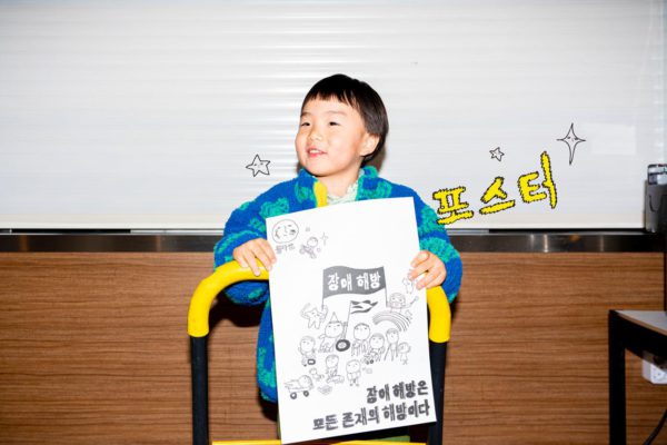 수레에 올라타 들다방 장애 해방 포스터를 들고 신난 아​​기.