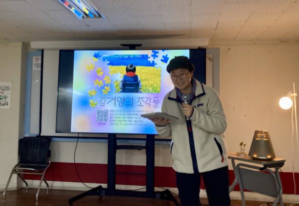 은은한 조명이 밝혀진 무대 커다란 액정화면에 김기영의 조각들 포스터가 크게 출력되어 있다. 그 앞으로 민아영 감독이 나와 마이크를 든 채 웃으며 무언가 안내하고 있다.