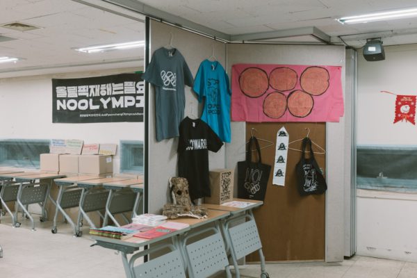 들다방 상영회장 뒤편 풍경. 벽면에 검정 바탕 흰 글씨로 노 올림픽스 현수막이 걸려 있고, 뒤편에 마련된 코너에 각종 올림픽반대 티셔츠와 가방, 플래그 등 굿즈 들이 전시돼 있다.