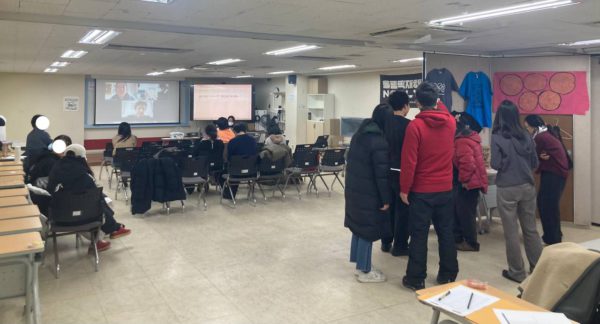 들다방 상영회장 쉬는 시간 풍경. 앞쪽 스크린에 화상회의 줌 미팅 창이 열려 있고, 의자에 앉은 사람들. 상영회장 뒤편에는 각종 올림픽반대 티셔츠와 가방, 플래그 등 굿즈 들이 전시돼 있다.