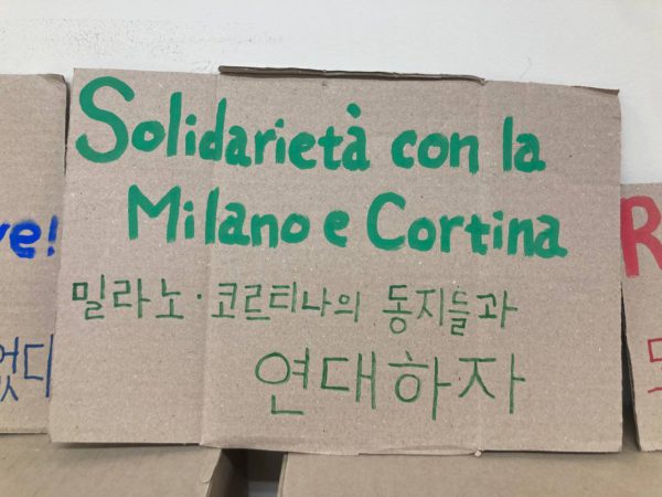 한글과 이탈리아어로 쓰인 초록 손피켓. 밀라노 코르티나의 동지들과 연대하자. Solidarità con la Milano e Cortina.