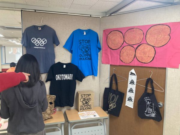 들다방 상영회장 뒤편 풍경. 각종 올림픽반대 티셔츠와 가방, 플래그 등 굿즈 들이 전시돼 있고 구경하는 사람들. 숭덩 베어진 통나무 줄기 다섯 개가 오륜기를 연상케 하는 빨간 깃발 아래 노 올림픽 검정 애코백 등이 전시된 모습.
