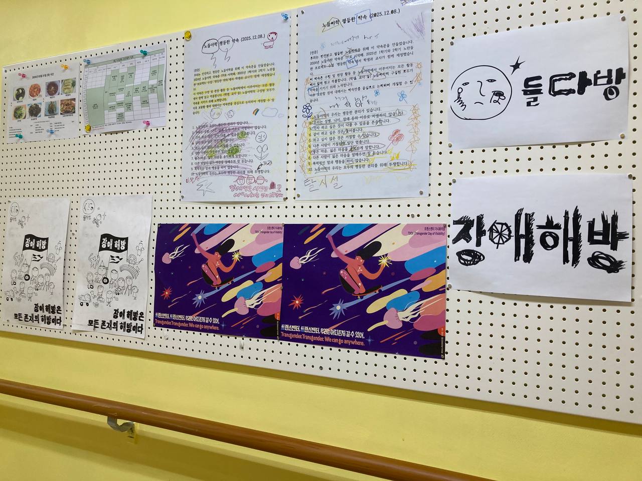들다방 복도 벽면을 멀리서 찍은 사진. 게시판에 들다방 장애해방 포스터와 이번 트랜스젠더 가시화의 날 포스터 촬영 사진이 나란히 붙어 있고, 그 외에도 들다방, 장애해방, 노들평등약속문, 식단표 등등이 옹기종이 붙어 있다. 