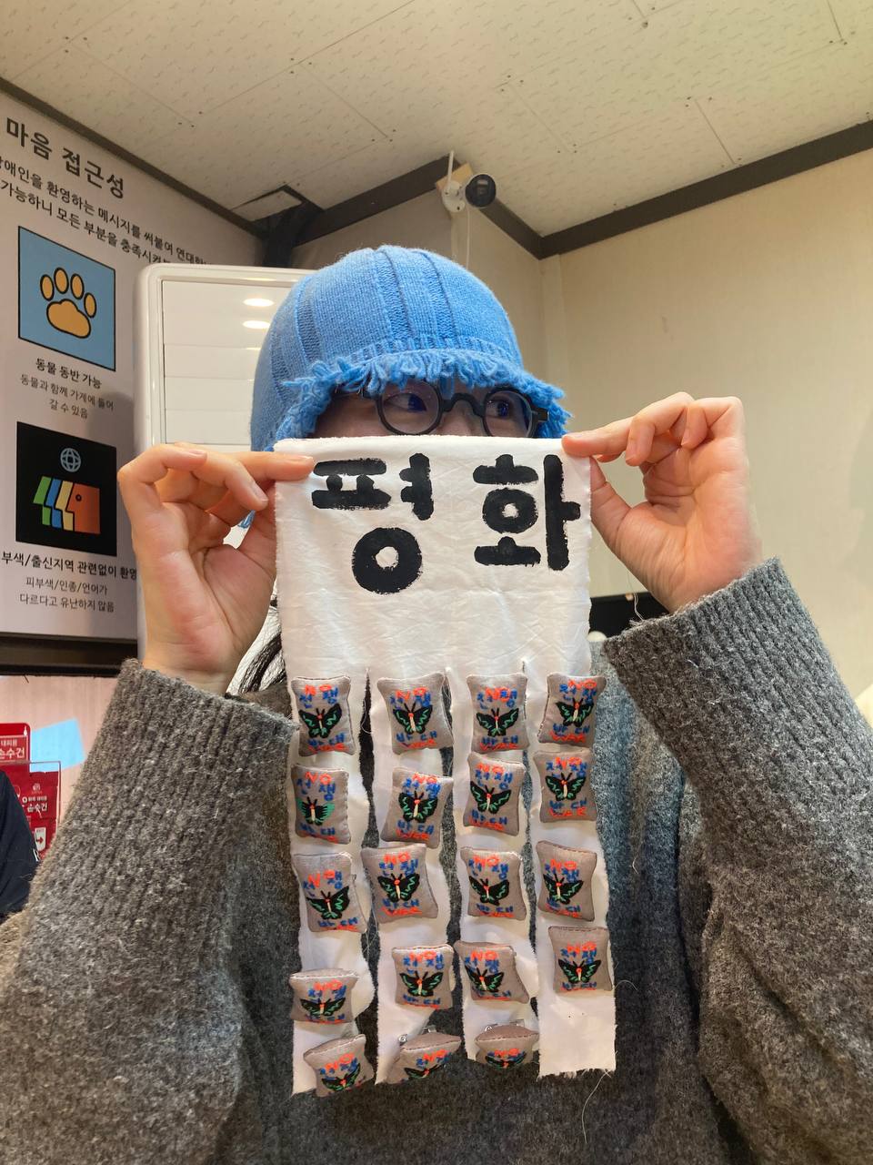 뱃지들이 달린 천을 들어 보이며 천 뒤에서 유희 작가가 눈을 빼꼼히 내보인 다른 각도 사진. 평화를 바라는 수공예 뱃지를 든 유희 작가님과 뱃지 약 20개 주렁주렁